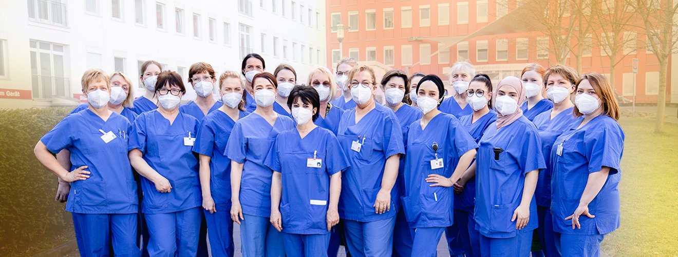 25 Hebammen in blauen Kitteln und FFP2-Maske stehen vor einem Krankenhaus