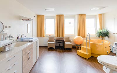 Kreißsaalraum mit gelber Badewanne, gelben Ball, zwei Stühlen und einen Tisch, drei Fenster, Waschbecken und Waage