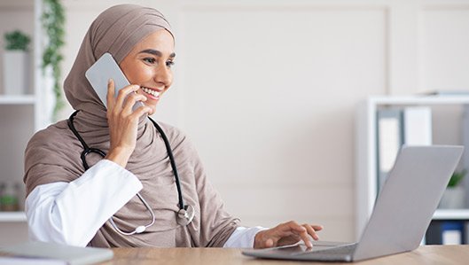 Eine Ärztin mit Kopftuch telefoniert und bedient ein Notebook