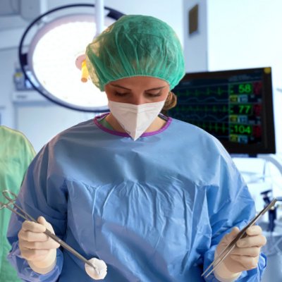 Pflegefachkraft mit blauem Kittel und güner OP-Haube steht im Operationssaal. In den Händen hält sie einen Tupfer und eine Pinzette.