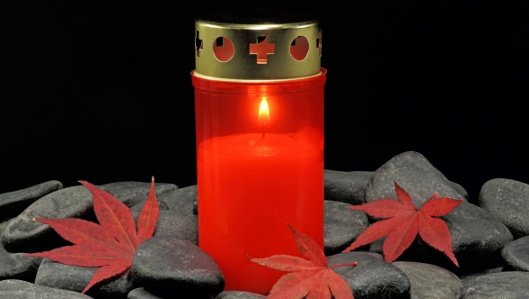 Symbolbild: ein brennendes rotes Grablicht auf grauen Kieselsteinen.