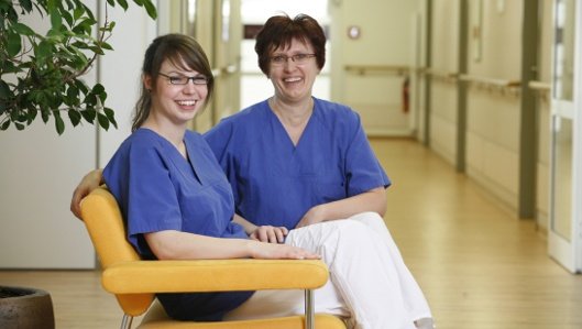 Symbolbild: Zwei Pflegekräfte auf einer Couch im Wartebereich lächeln in die Kamera.