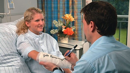 Ein Arzt schreibt mit einem Filzstift  -Gute Besserung- auf den Unterarm-Gips einer Patientin, die in ihrem Bett liegt. 