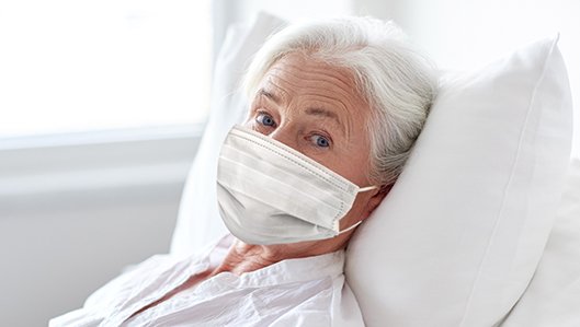 Eine alte Dame liegt im Krankenbett und trägt eine medizinische Maske.