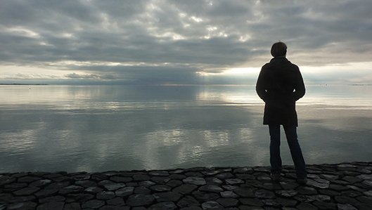Symbolbild: Ein Mann schaut gedankenverloren auf einen See, bewölkter Abendhimmel. 