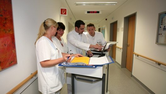 Symbolbild: Zwei Ärzte und zwei Pflegekräfte bespechen einen Befund auf dem Krankenhausflur am Visitenwagen.